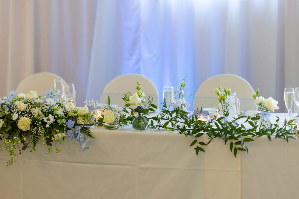 A decorated table with white linen, green foliage, white and light blue flowers, candles, and glassware against a white curtain backdrop. Copyright Somerside Photography Ltd // www.somersidephotography.com