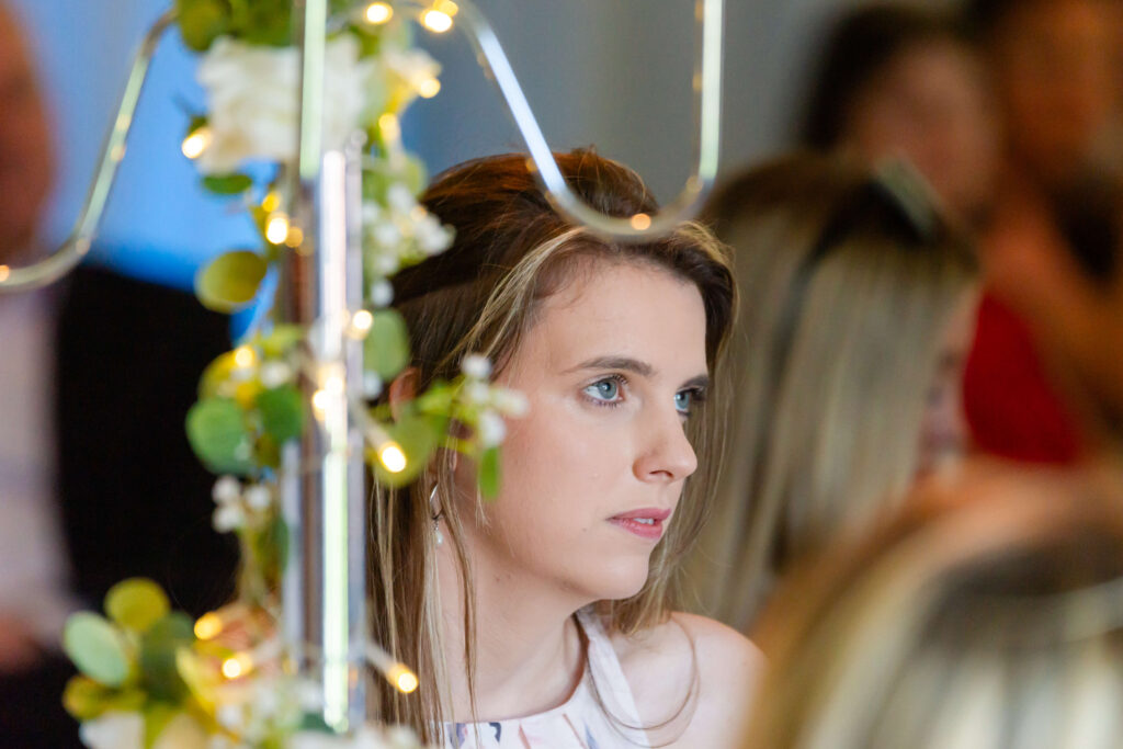 A young woman with light brown hair sits indoors, looking attentively ahead. Decorative lights and flowers are in the foreground, with blurred people in the background. Copyright Somerside Photography Ltd // www.somersidephotography.com