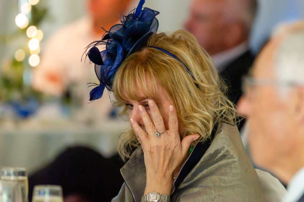A woman wearing a blue fascinator covers part of her face with her hand, appearing emotional at a formal event. Copyright Somerside Photography Ltd // www.somersidephotography.com