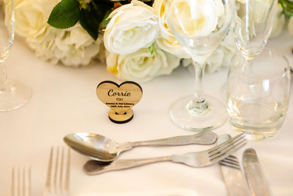 A wooden heart-shaped name card reading "Carrie" is placed on a white table set with cutlery, glassware, and white floral arrangements. Copyright Somerside Photography Ltd // www.somersidephotography.com