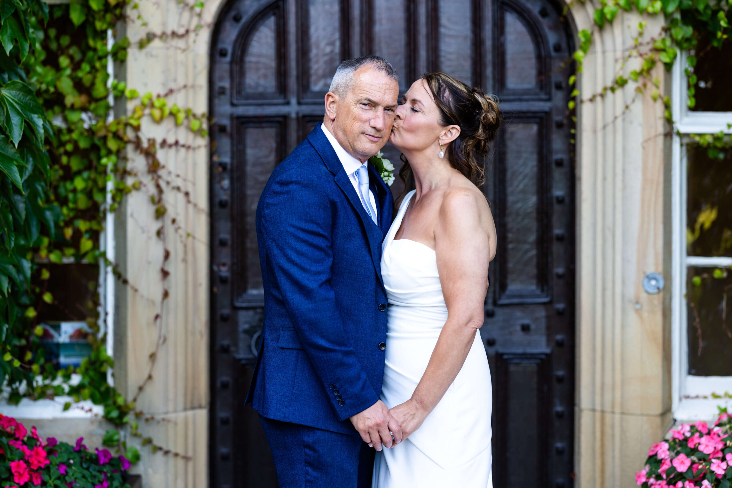 A couple in formal wedding attire stand holding hands in front of a large wooden door; the woman kisses the man on the cheek. Copyright Somerside Photography Ltd // www.somersidephotography.com