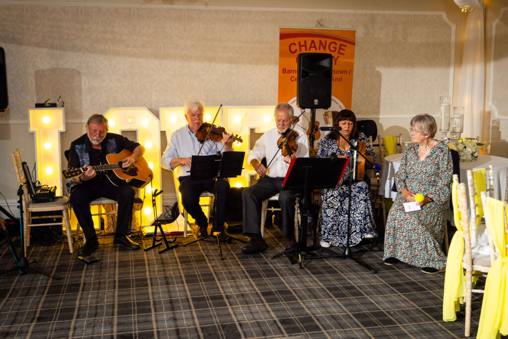 Five musicians, seated in a row, perform with guitars, violins, and a flute. A sign behind them reads "CHANGE". The room is decorated with yellow and white accents. Copyright Somerside Photography Ltd // www.somersidephotography.com