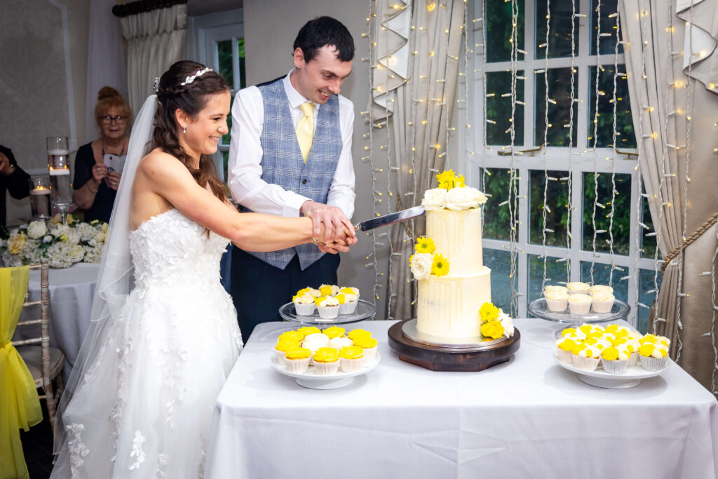 A bride and groom smile as they cut a white tiered wedding cake decorated with yellow flowers, surrounded by cupcakes, inside a decorated venue. Copyright Somerside Photography Ltd // www.somersidephotography.com