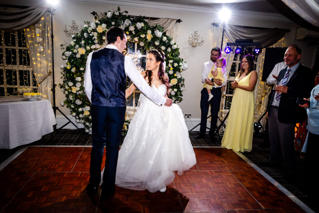 A bride and groom share their first dance in front of a floral arch, whilst guests watch and smile in a decorated reception venue. Copyright Somerside Photography Ltd // www.somersidephotography.com