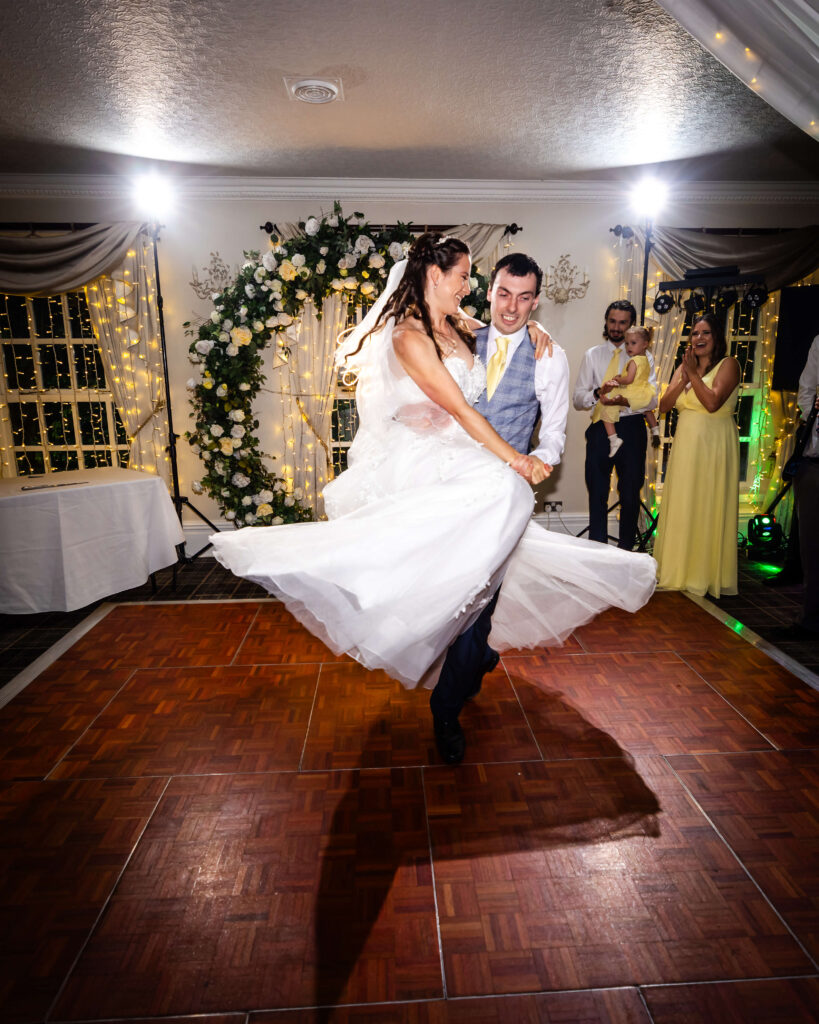 A bride and groom dance on a wooden floor in a decorated room, with the bride’s dress twirling and guests watching in the background. Copyright Somerside Photography Ltd // www.somersidephotography.com