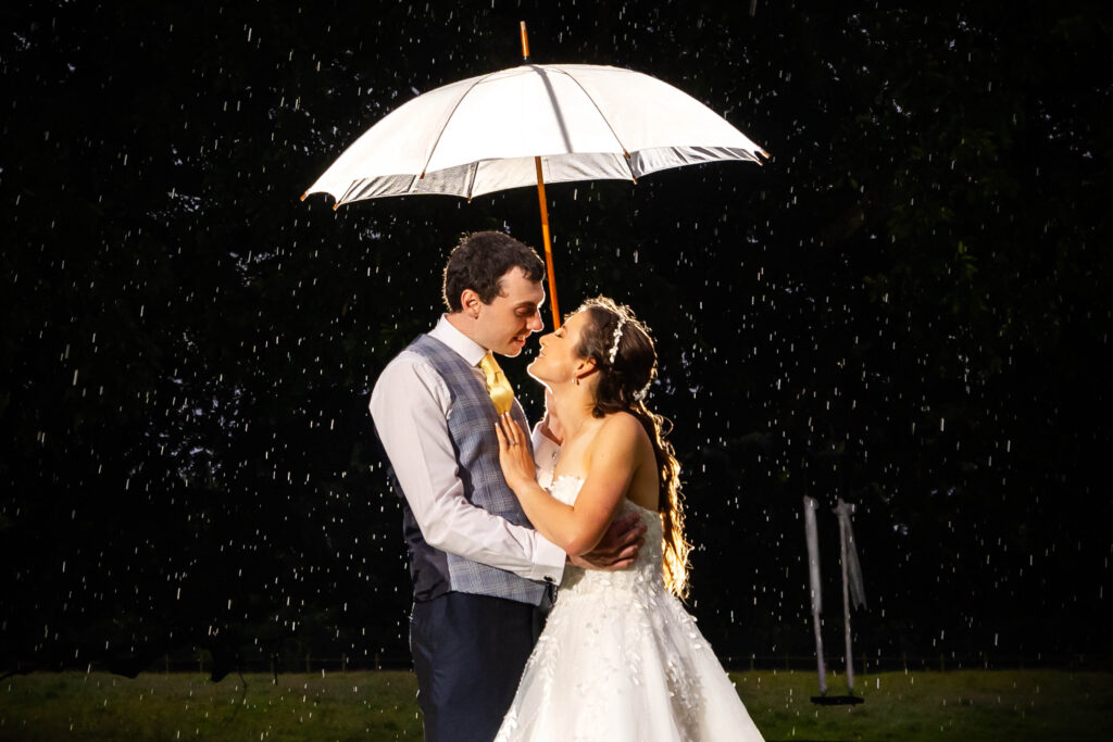 A bride and groom stand under a white umbrella in the rain at night, looking at each other and smiling. Copyright Somerside Photography Ltd // www.somersidephotography.com