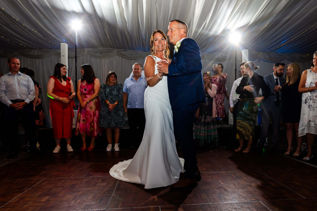 A bride and groom share their first dance in a decorated venue, surrounded by guests watching and standing in a semicircle. Copyright Somerside Photography Ltd // www.somersidephotography.com
