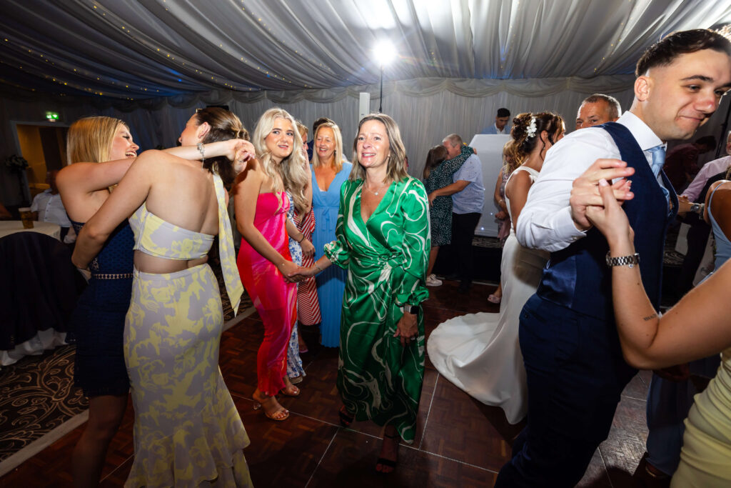A group of people dressed in formal attire dance and smile together at an indoor event under draped ceiling décor. Copyright Somerside Photography Ltd // www.somersidephotography.com
