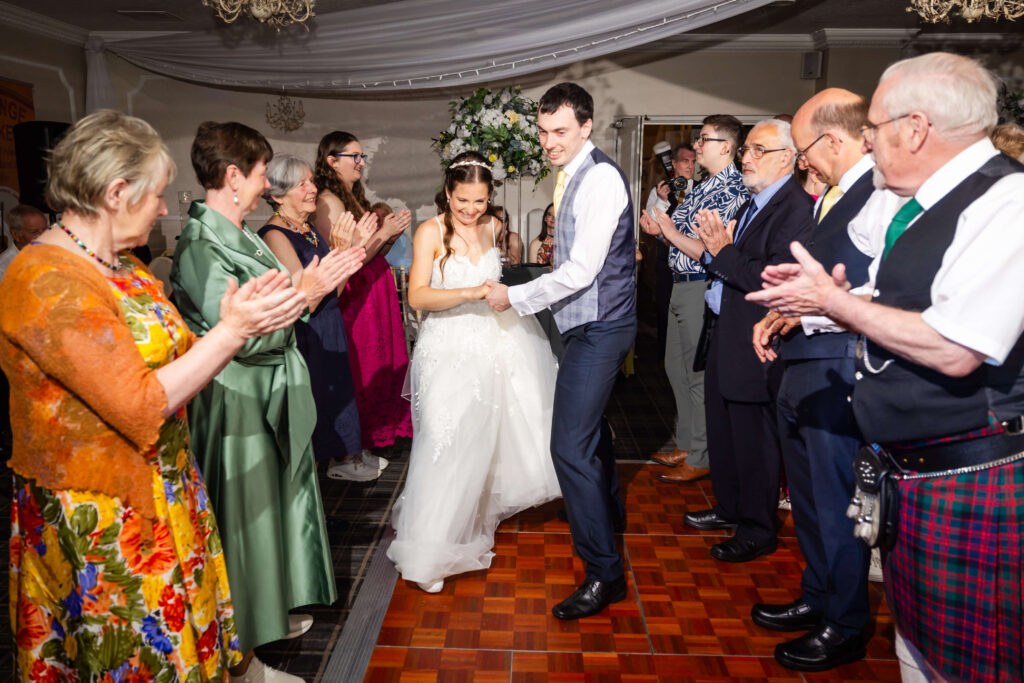 A newlywed couple dances together while guests stand on either side, clapping and smiling in a brightly lit indoor venue. Copyright Somerside Photography Ltd // www.somersidephotography.com