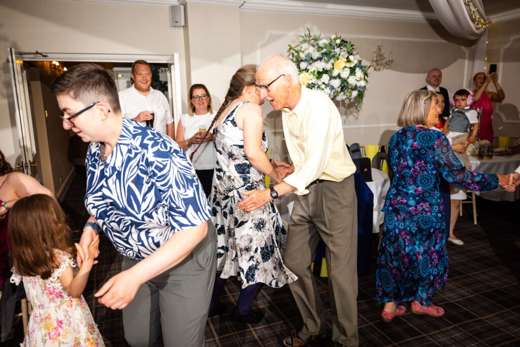A group of people of various ages are dancing together in a brightly lit room decorated with flowers and tables. Copyright Somerside Photography Ltd // www.somersidephotography.com