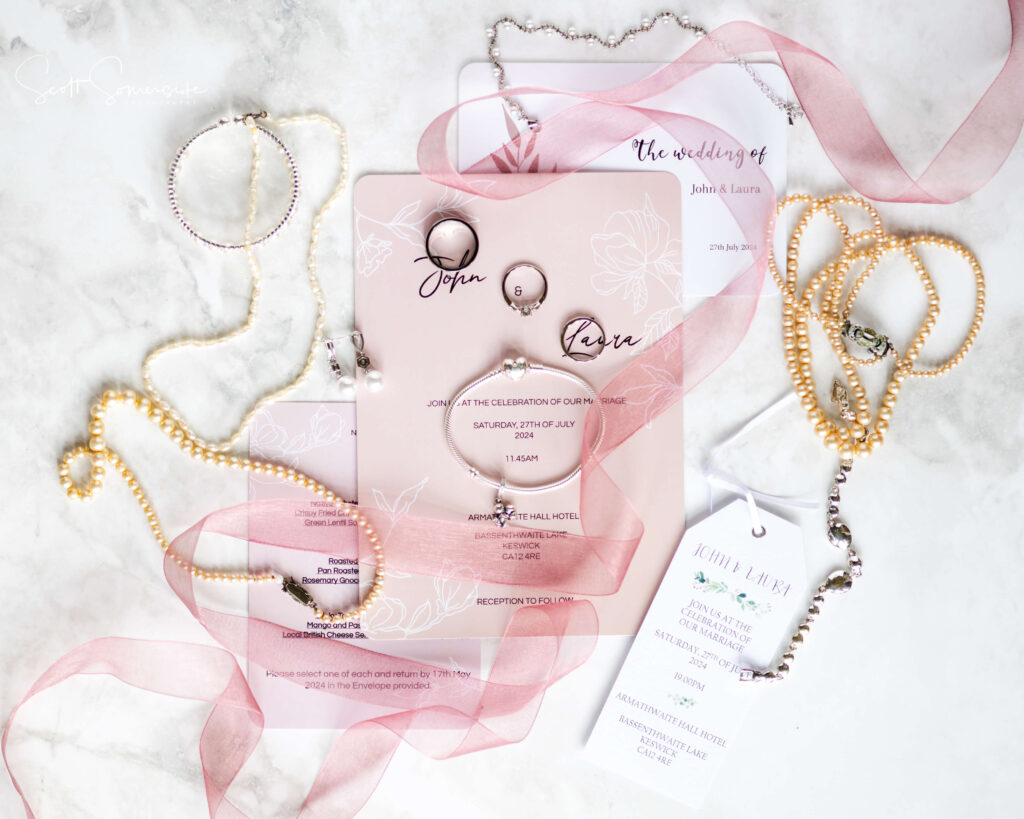 Flat lay of wedding invitations, pink ribbon, pearl and silver jewellery, and a wedding ring on a marble surface. Copyright Somerside Photography Ltd // www.somersidephotography.com