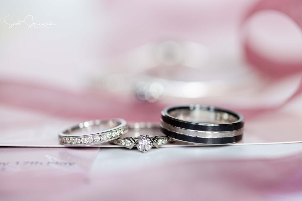 Three rings, including a diamond engagement ring and two wedding bands, are displayed on a white and pink surface with a blurred background. Copyright Somerside Photography Ltd // www.somersidephotography.com