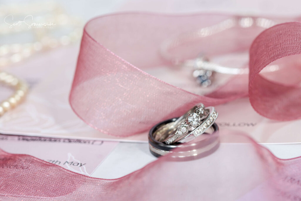 Close-up of two silver rings with diamonds placed on a pink ribbon, with pearl necklace and soft pink background elements. Copyright Somerside Photography Ltd // www.somersidephotography.com