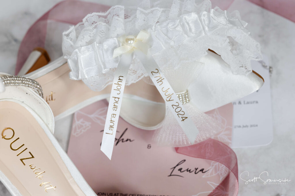 A white bridal shoe and lace garter with a ribbon reading "Laura and John 27th July 2024" are arranged on a pink invitation card. Copyright Somerside Photography Ltd // www.somersidephotography.com