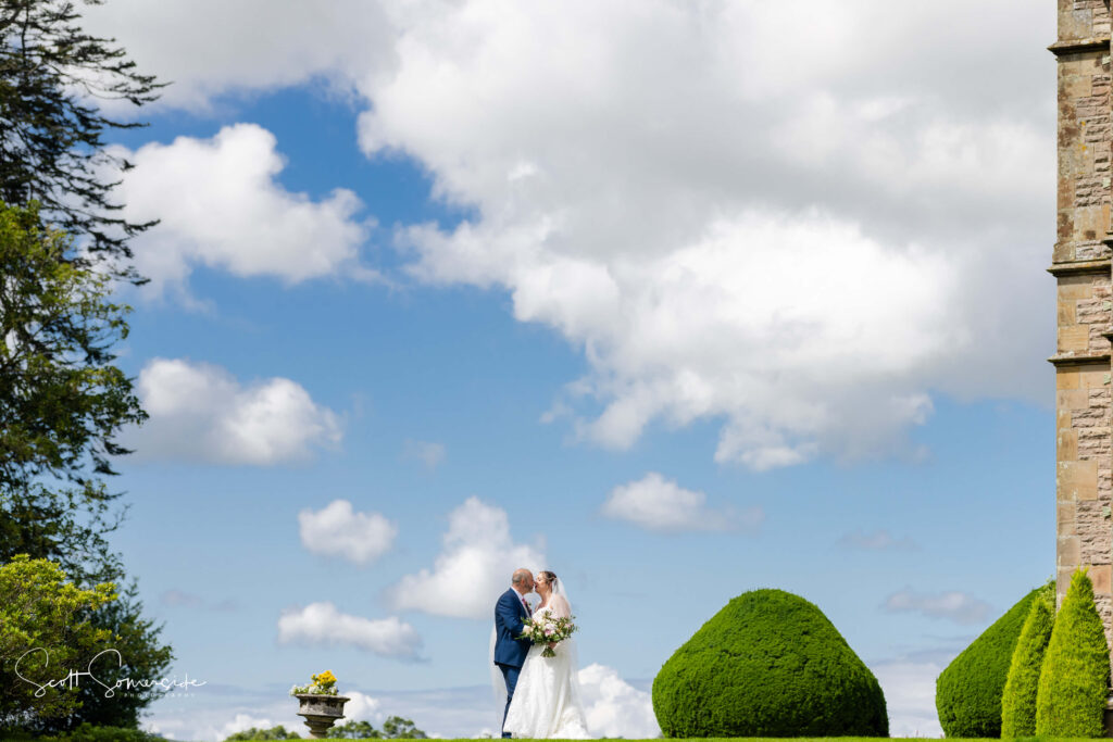 A bride and groom stand together outdoors, surrounded by greenery and under a partly cloudy sky, with a stone building partially visible on the right. Copyright Somerside Photography Ltd // www.somersidephotography.com