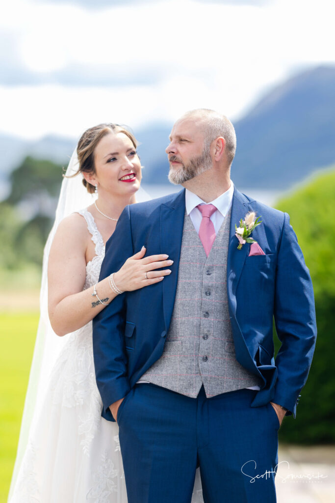 A bride in a white dress stands behind a groom in a blue suit and pink tie outdoors, with mountains and greenery in the background. Copyright Somerside Photography Ltd // www.somersidephotography.com
