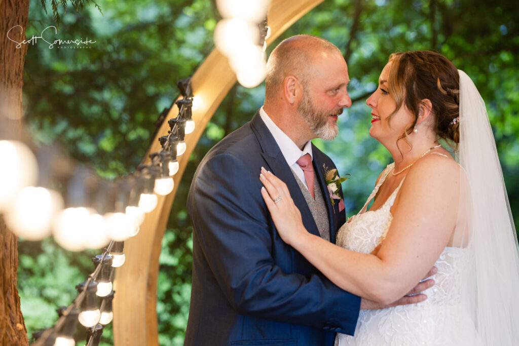 A bride and groom stand close together, smiling at each other, with fairy lights and greenery in the background. Copyright Somerside Photography Ltd // www.somersidephotography.com