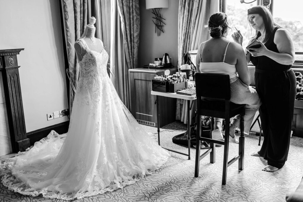 A bride sits by a window having her make-up done while her wedding dress hangs on a mannequin nearby in a well-lit room. Copyright Somerside Photography Ltd // www.somersidephotography.com