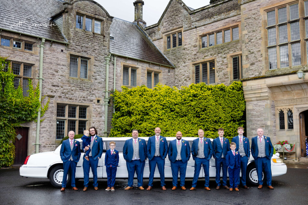 A group of men and two boys in blue suits stand in front of a white limousine, posing outside a large stone building covered in greenery. Copyright Somerside Photography Ltd // www.somersidephotography.com