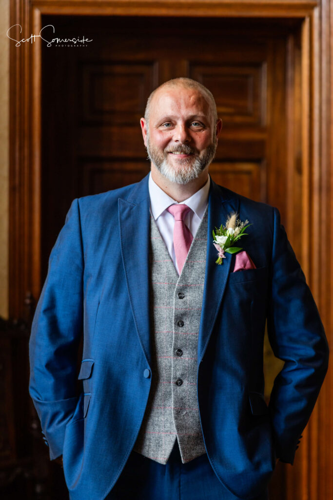 A man with a beard in a blue suit, pink tie, and buttonhole stands indoors in front of a wooden door, smiling with his hands in his pockets. Copyright Somerside Photography Ltd // www.somersidephotography.com