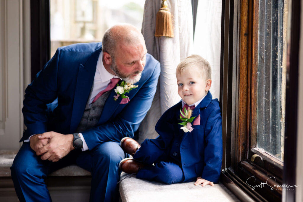An older man in a blue suit sits beside a young boy in a matching suit by a window. Both wear buttonholes and look relaxed. Copyright Somerside Photography Ltd // www.somersidephotography.com