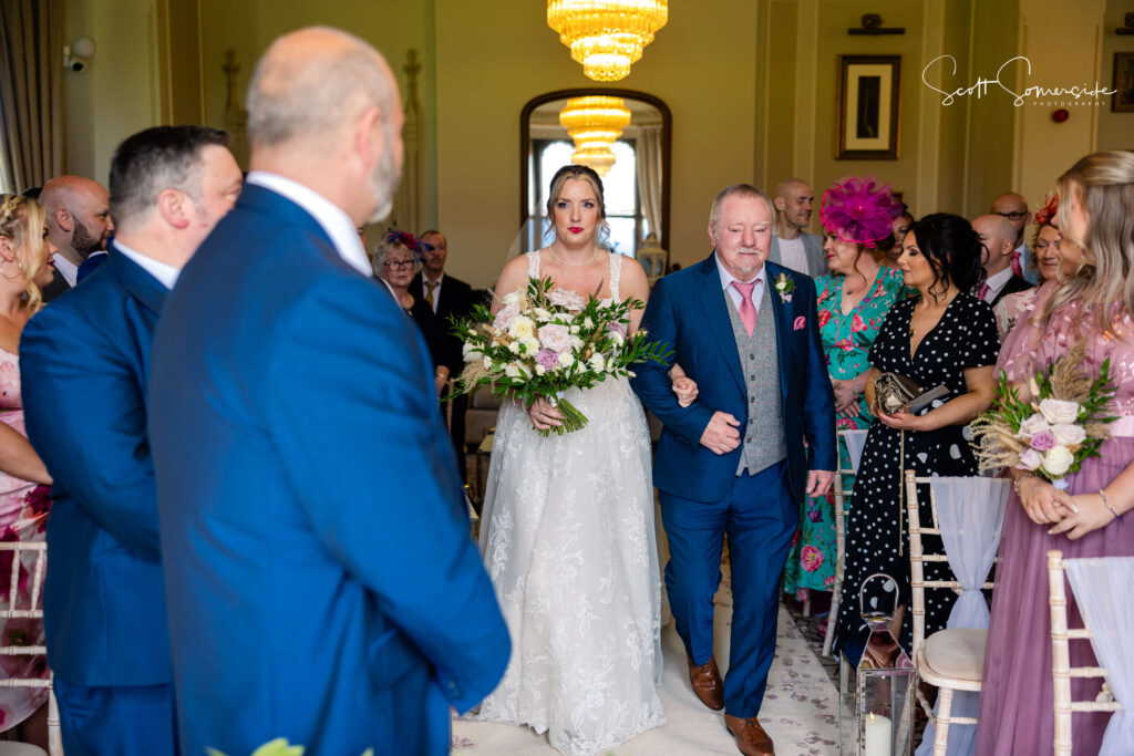 A bride in a white dress walks down the aisle with an older man, whilst guests seated on either side watch in a decorated indoor venue. Copyright Somerside Photography Ltd // www.somersidephotography.com