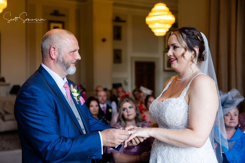 A bride and groom exchange rings during their wedding ceremony in an indoor venue, with guests seated in the background. Copyright Somerside Photography Ltd // www.somersidephotography.com