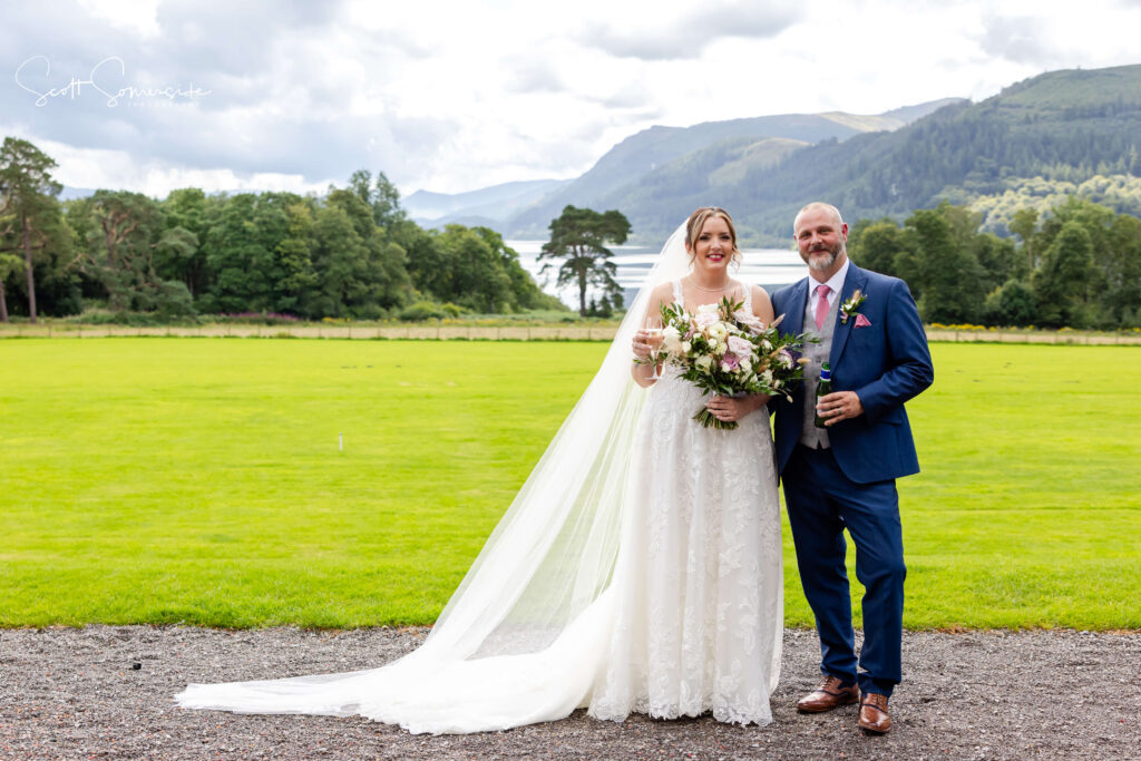Bride in a white wedding dress and veil stands beside a man in a blue suit holding drinks, with a bouquet in hand, on a lawn with mountains and a lake in the background. Copyright Somerside Photography Ltd // www.somersidephotography.com