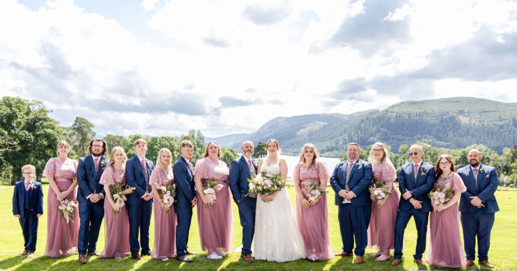 A wedding party of fourteen adults and one child, dressed in formal attire, stands in a row outdoors with mountains and greenery in the background. Copyright Somerside Photography Ltd // www.somersidephotography.com