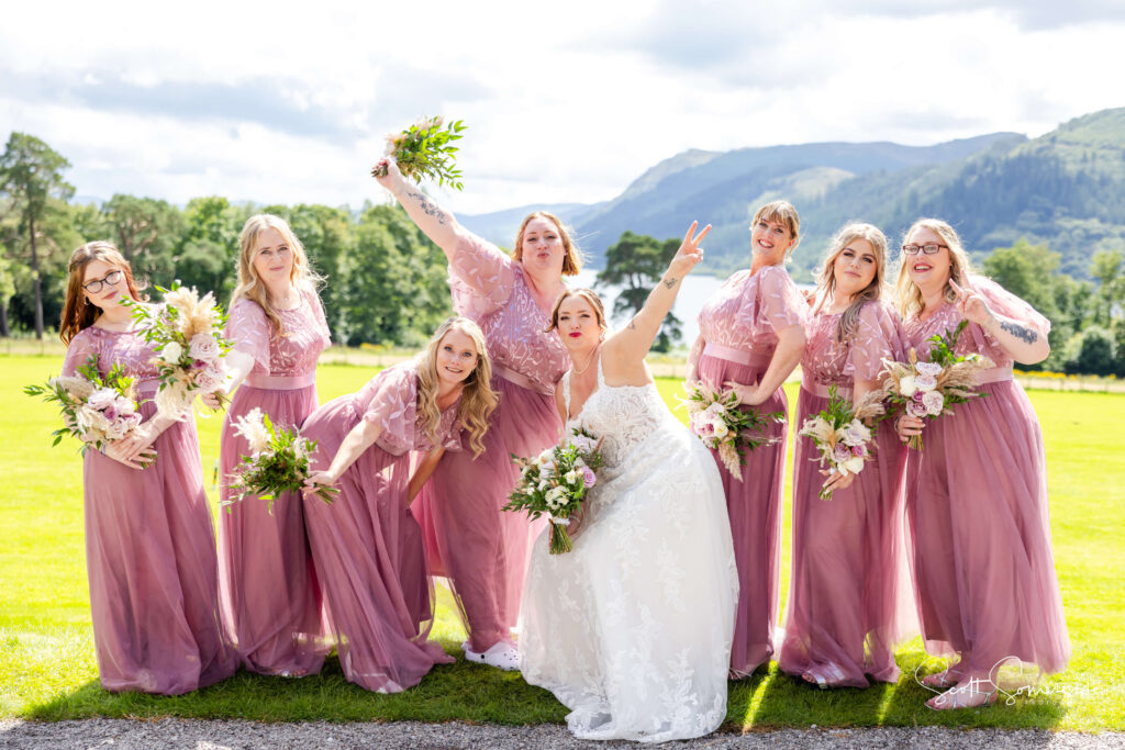 A bride in a white dress poses with seven bridesmaids in matching mauve dresses holding bouquets, standing outdoors on a sunny day with mountains in the background. Copyright Somerside Photography Ltd // www.somersidephotography.com
