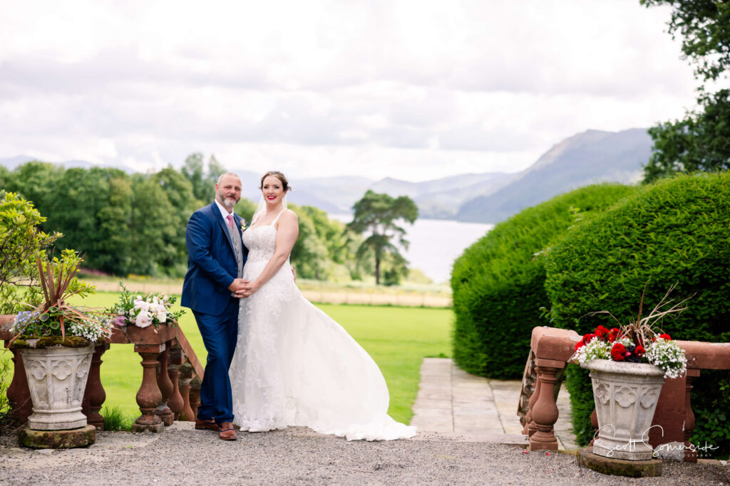 A bride and groom stand together outdoors, smiling, with green gardens, stone planters, and a lake with mountains in the background. Copyright Somerside Photography Ltd // www.somersidephotography.com