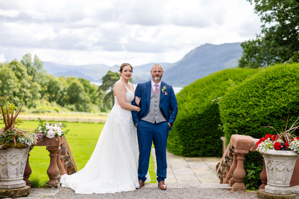 A bride in a white wedding dress and a groom in a blue suit stand together outdoors on a stone path, with greenery and mountains in the background. Copyright Somerside Photography Ltd // www.somersidephotography.com