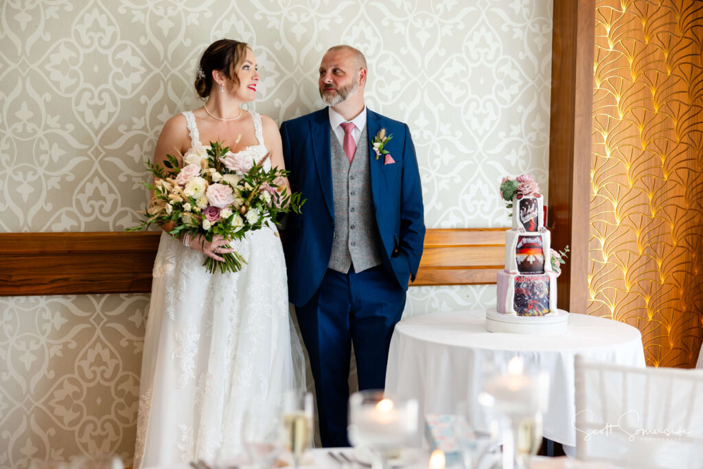 A bride holding a bouquet and a groom in a blue suit stand side by side, smiling at each other beside a table with a tiered wedding cake. Copyright Somerside Photography Ltd // www.somersidephotography.com