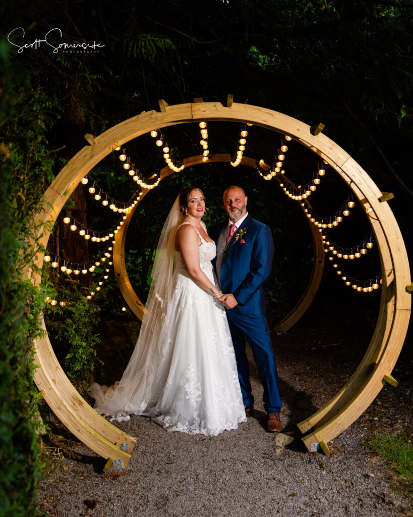 Bride and groom stand together under a circular wooden arch decorated with fairy lights, surrounded by greenery, at night. Copyright Somerside Photography Ltd // www.somersidephotography.com