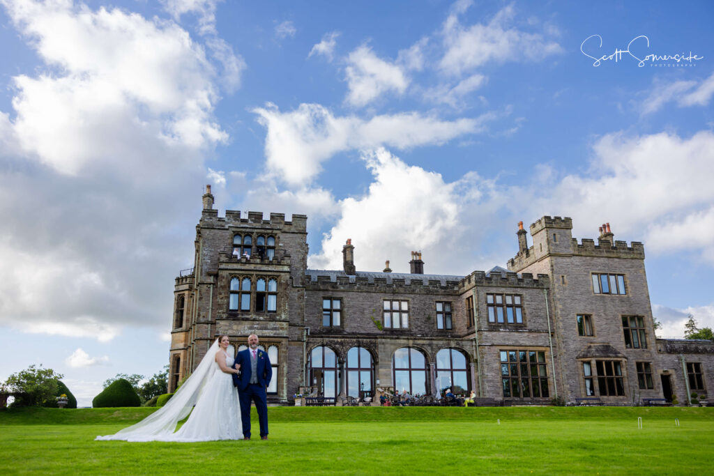A bride and groom stand on a green lawn in front of a large stone castle-like building under a partly cloudy sky. Copyright Somerside Photography Ltd // www.somersidephotography.com