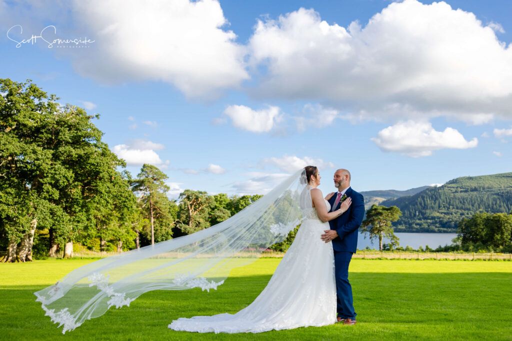 A bride and groom stand together outdoors on a grassy field, with the bride's veil flowing in the wind and trees, hills, and a lake in the background under a blue sky. Copyright Somerside Photography Ltd // www.somersidephotography.com