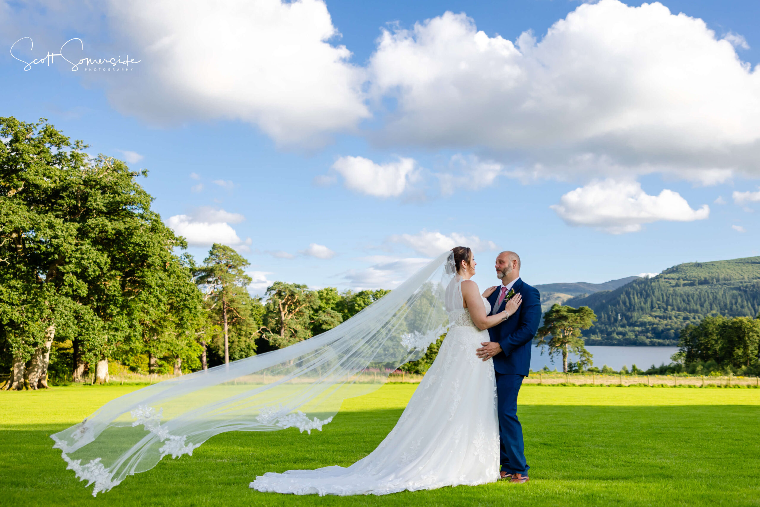 A bride and groom stand together outdoors on a grassy field, with the bride's veil flowing in the wind and trees, hills, and a lake in the background under a blue sky. Copyright Somerside Photography Ltd // www.somersidephotography.com