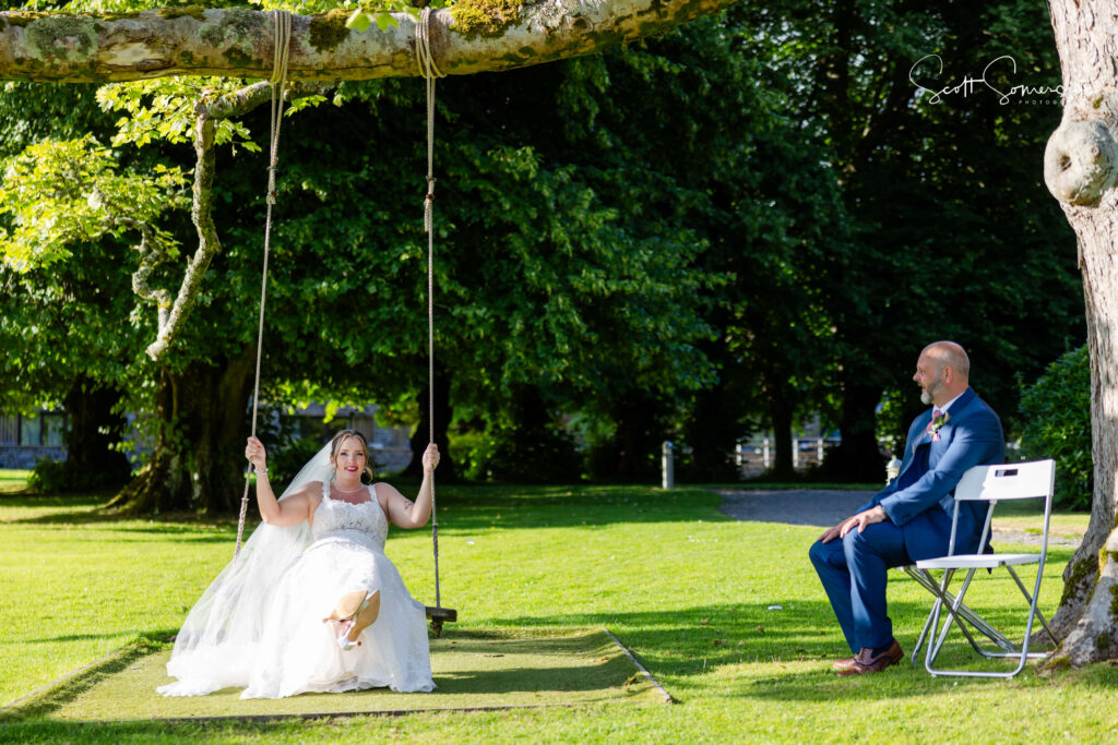 A bride in a white dress and veil sits on a tree swing while a man in a blue suit watches her from a chair on a sunny garden. Copyright Somerside Photography Ltd // www.somersidephotography.com