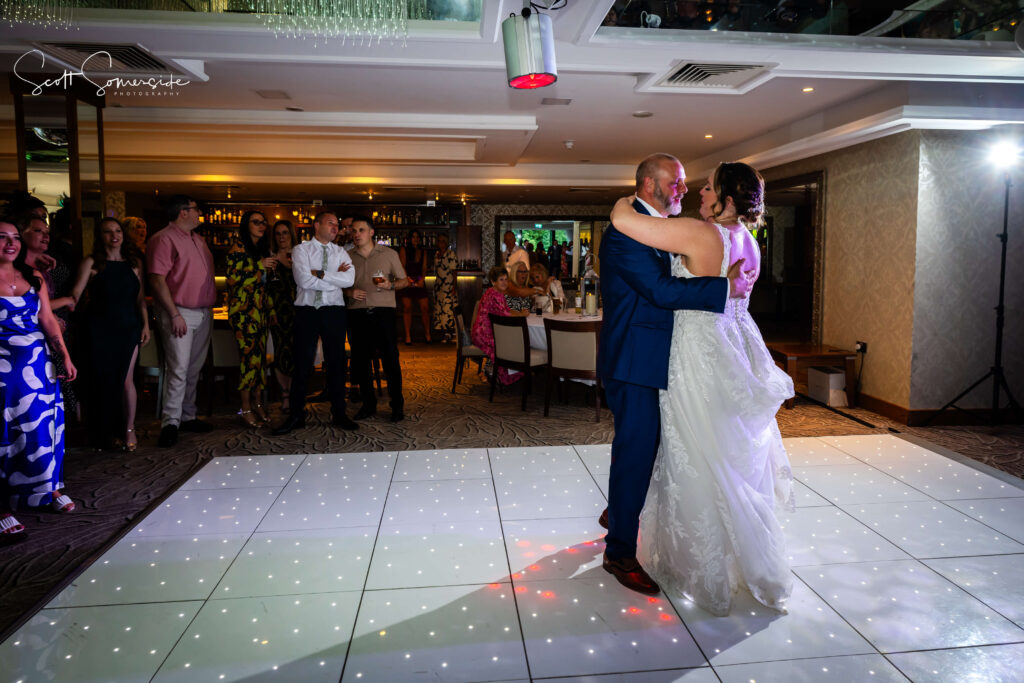 A bride and groom share their first dance on a lit-up dance floor while wedding guests watch and smile in a banqueting hall. Copyright Somerside Photography Ltd // www.somersidephotography.com