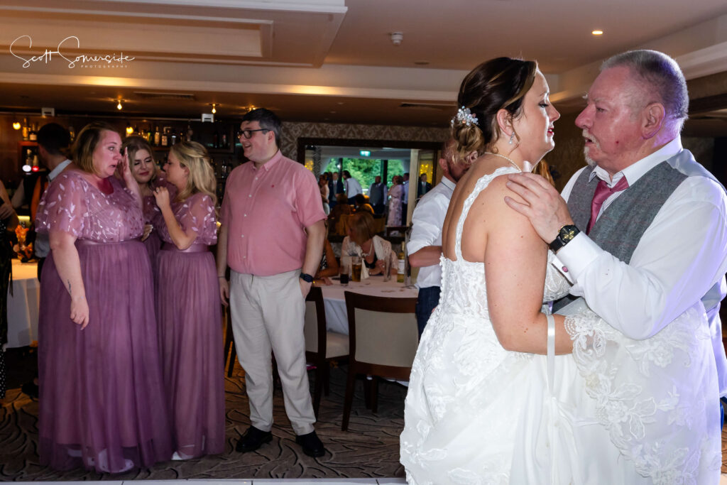 A bride in a white dress dances with an older man, while guests in pink and purple outfits talk in the background at a wedding reception. Copyright Somerside Photography Ltd // www.somersidephotography.com