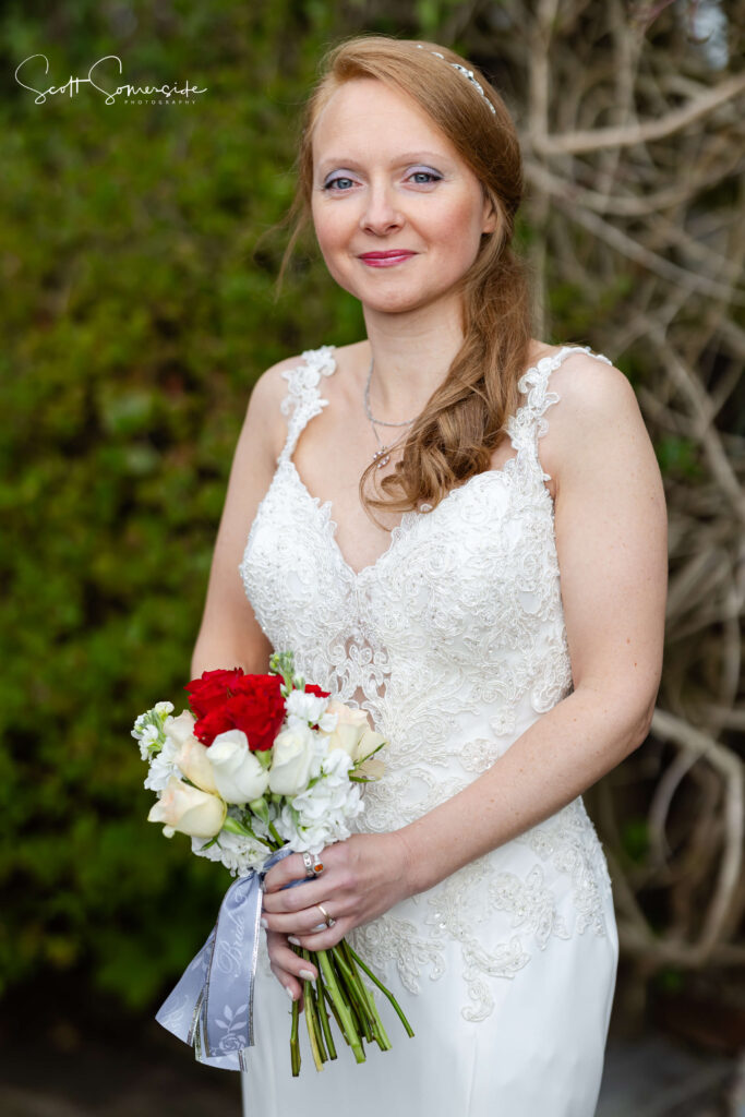 A woman in a white lace wedding dress holds a bouquet of red and white flowers, posing outdoors in front of greenery and bare branches. Copyright Somerside Photography Ltd // www.somersidephotography.com