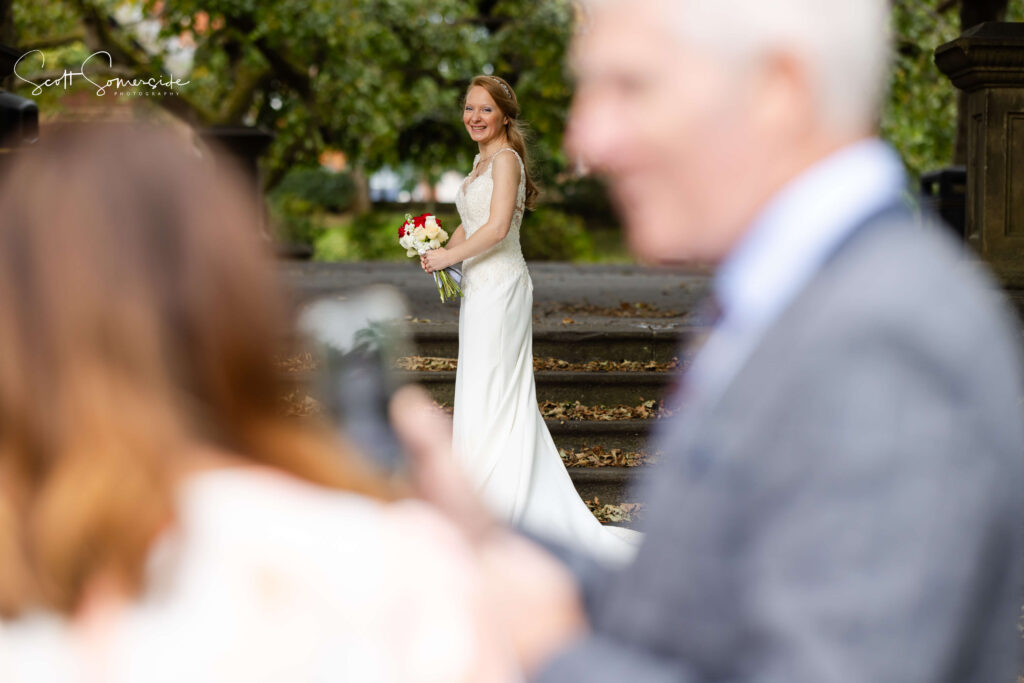 A bride in a white dress holding a bouquet stands on stone steps outdoors, smiling; two people are blurred in the foreground. Copyright Somerside Photography Ltd // www.somersidephotography.com