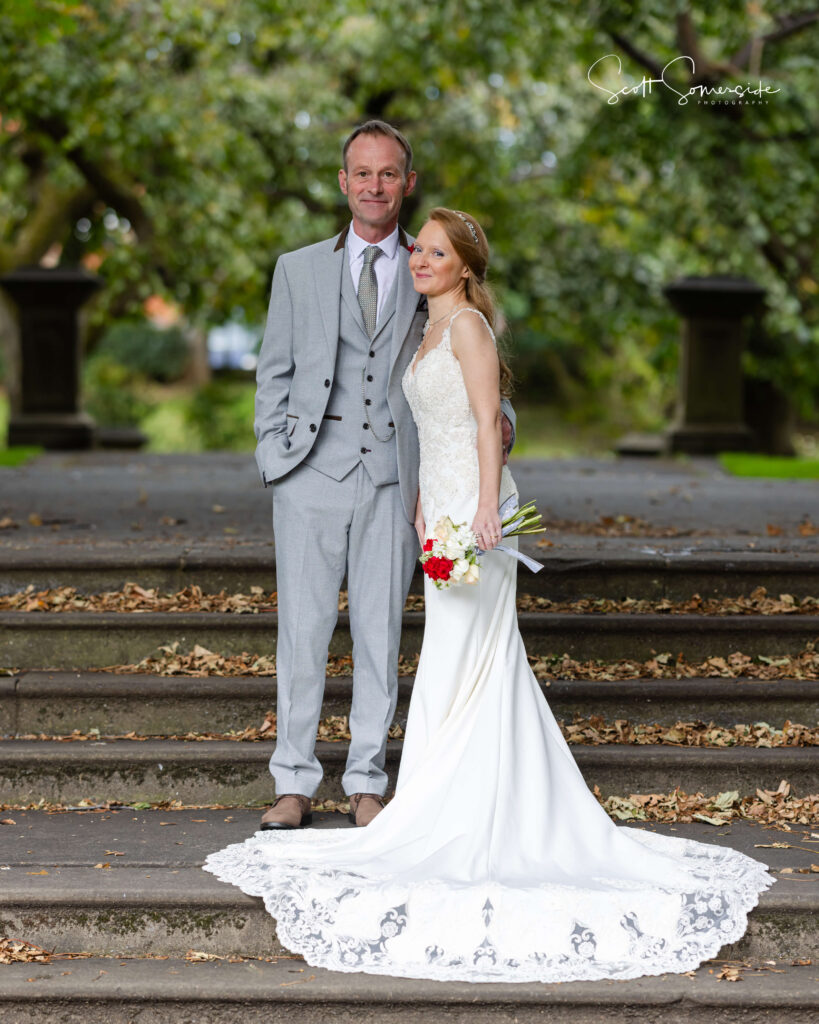 A bride in a white dress and a groom in a light grey suit stand together on outdoor stone steps, surrounded by trees and fallen leaves. Copyright Somerside Photography Ltd // www.somersidephotography.com