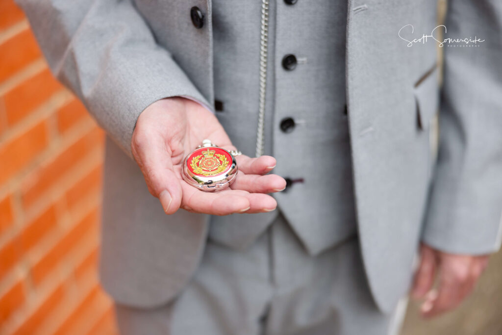 A person in a light grey suit holds an ornate pocket watch with a red and gold design in their left hand. Copyright Somerside Photography Ltd // www.somersidephotography.com