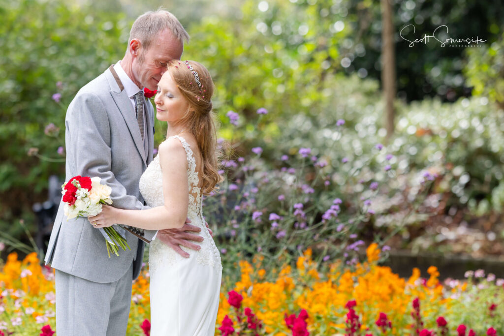 A bride and groom stand close together in a garden, surrounded by colourful flowers, as the groom gently holds the bride and she holds a bouquet. Copyright Somerside Photography Ltd // www.somersidephotography.com