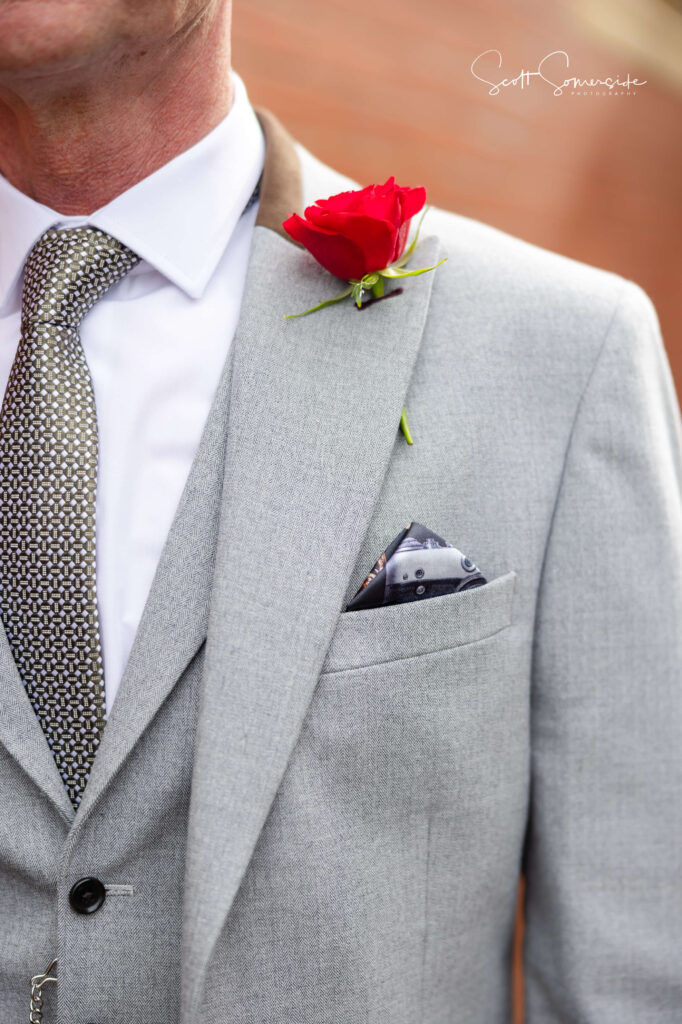 A man wearing a light grey suit, white shirt, patterned tie, and a red rose buttonhole in his lapel pocket. Copyright Somerside Photography Ltd // www.somersidephotography.com