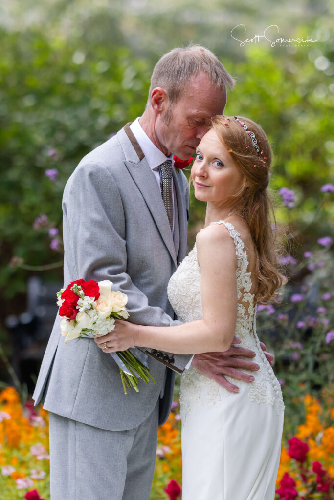 A bride in a white dress holding a bouquet stands in front of a groom in a grey suit who is kissing her forehead in a garden with colourful flowers. Copyright Somerside Photography Ltd // www.somersidephotography.com