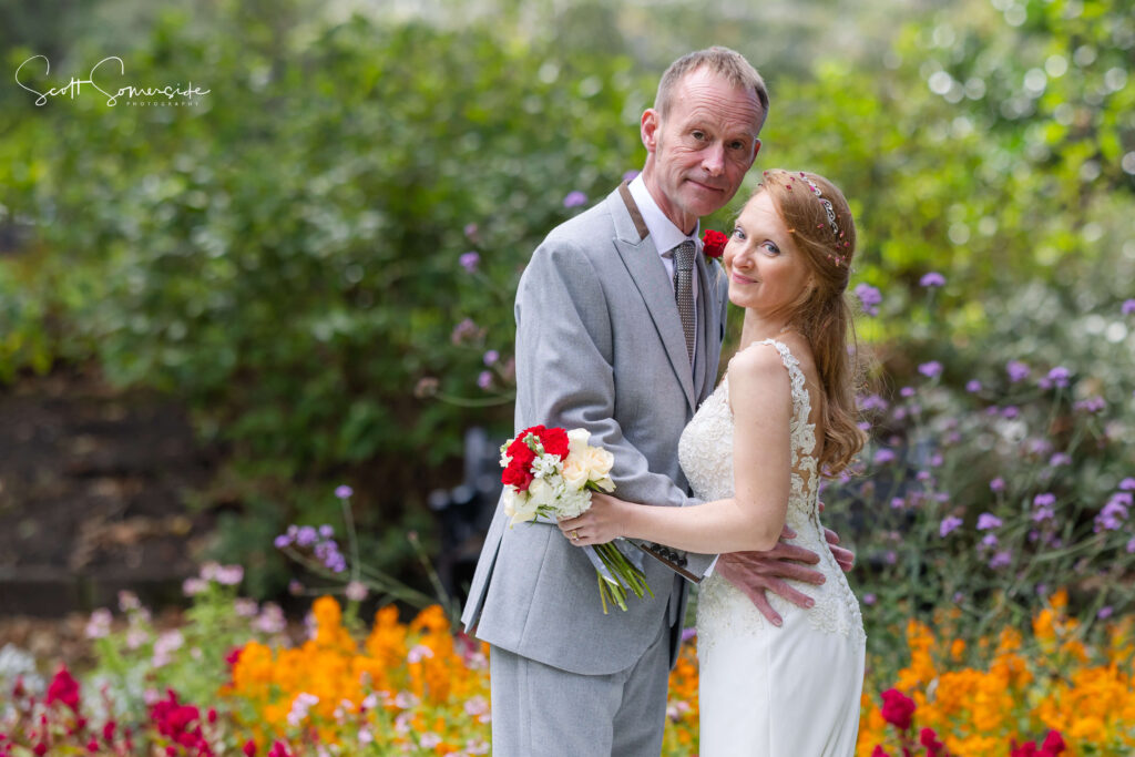 A bride and groom stand close together in a garden, surrounded by colourful flowers; the bride holds a bouquet and the groom wears a grey suit. Copyright Somerside Photography Ltd // www.somersidephotography.com