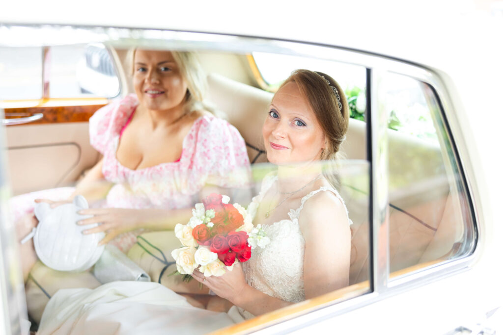 A bride in a white dress holding a bouquet sits next to a bridesmaid in pink inside a car, both looking out of the window. Copyright Somerside Photography Ltd // www.somersidephotography.com
