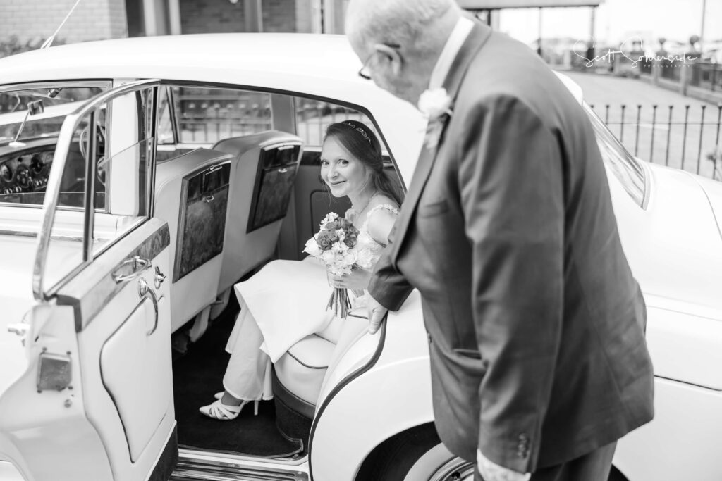 A bride holding a bouquet sits in the back seat of a car whilst an older man stands by the open door, looking at her. Copyright Somerside Photography Ltd // www.somersidephotography.com